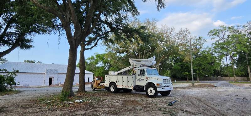 White bucket truck with its boom folded, parked under two live oaks on a gravel lot beside a building in house wrap