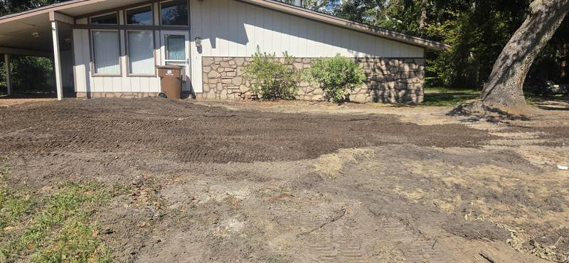Freshly graded bare-dirt yard with track marks in front of a white ranch house, one large oak left standing at the edge