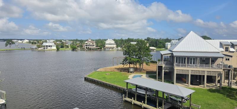 View from high up over a wide bayou: a raised gray house with a metal roof, a boathouse with two boats and a pool
